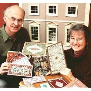 Janet and Chris in front of Janet's dollshouse, holding some of her needlepoint carpets